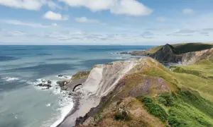 Grüne Hügel und steile Klippen am South West Coast Path. Das Meer glitzert in der Sonne, Wolken ziehen über den blauen Himmel.