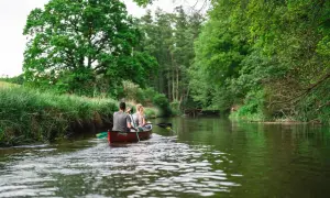 Zwei Personen paddeln in einem Kanu durch einen Fluss, umgeben von üppiger grüner Natur. Ein friedlicher Moment im **Warnow Durchbruchstal**.