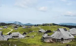 Idyllische Holzhütten auf grünen Hügeln, ein malerischer Ort für eine Velika Planina Wanderung. Berge im Hintergrund.