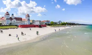 Ein sonniger Ostsee Strand mit Menschen, die spazieren gehen und Kinder spielen. Im Hintergrund ein prächtiges Gebäude im Bäderstil.