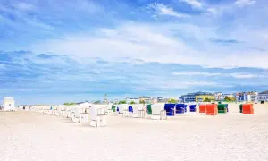 Strandkörbe in bunten Farben säumen den Sandstrand eines Ostseebads in Mecklenburg-Vorpommern. Im Hintergrund die Promenade mit Häusern und ein Leuchtturm.