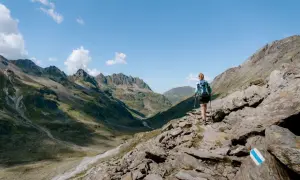 Frau mit Rucksack und Stöcken wandert auf felsigem Pfad im Montafon. Bergpanorama mit Tälern und Gipfeln.