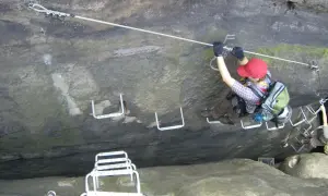 Ein Klettersteig im Elbsandsteingebirge: Eine Person sichert sich an einem Stahlseil, während sie über Felsstufen klettert.