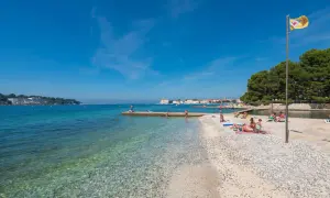 Sonniger istrien strand mit klarem Wasser, Kiesstrand und Badegästen. Im Hintergrund eine Stadtkulisse und grüne Bäume.