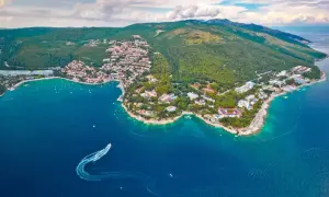 Blick auf eine Küstenstadt mit roten Dächern und grünen Hügeln, umgeben von türkisfarbenem Wasser. Ein Boot hinterlässt eine Spur im Meer. Ein echter **Istrien Geheimtipp**.