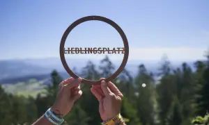Hände halten ein Schild mit "LIEBLINGSPLATZ" vor einer malerischen Bergkulisse. Ein echter Geheimtipp im Bayerischen Wald.