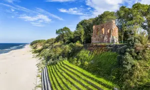 Ruinen einer alten Kirche thronen über einem Strand an der polnischen Ostsee. Terrassierte grüne Hänge führen zum Meer.