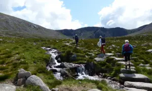 Drei Wanderer genießen das Cairngorms National Park Wandern entlang eines steinigen Pfades mit einem plätschernden Bach.