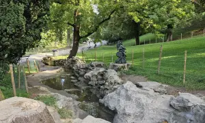 Ein kleiner Bach schlängelt sich durch den Parc des Buttes Chaumont, vorbei an Felsen und einer Skulptur. Grüne Hänge und Bäume säumen den Weg.