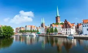 Historische Gebäude und Kirchtürme säumen das Wasser in Lübeck, einem der schönsten Ostsee Orte. Boote liegen am Kai, die Sonne scheint.