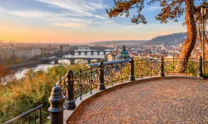 Prag im Herbst: Blick über die Moldau mit Brücken und Stadtkulisse bei Sonnenuntergang. Ein malerischer Ausblick von einem Aussichtspunkt mit gepflastertem Boden und schmiedeeisernem Geländer.