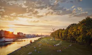 Sonnenuntergang an der Neckarwiese Mannheim: Menschen entspannen auf der Wiese, ein Schiff fährt auf dem Fluss, im Hintergrund Industrie und Bäume.