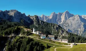 Alpe-Adria-Trail Erfahrungsbericht: Malerisches Bergdorf mit Kirche und Häusern, umgeben von Nadelwäldern und imposanten Gipfeln.