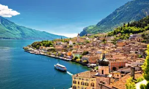 Buntes Dorf am Seeufer, Berge im Hintergrund. Ein Schiff fährt auf dem Wasser. Ein malerischer Ort am Lago Maggiore oder Gardasee.