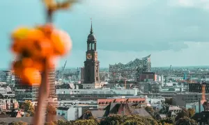 Blick auf Hamburg sehenswertes: Kirchturm St. Michaelis und Elbphilharmonie vor bewölktem Himmel. Im Vordergrund unscharfe orangefarbene Blüten.