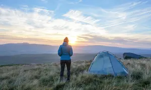 Frau blickt auf Sonnenuntergang, ihr Zelt steht bereit für das Wandern mit Zelt. Die Landschaft ist hügelig, der Himmel dramatisch.
