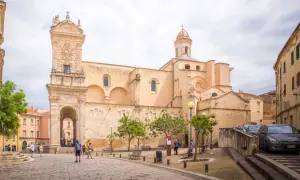 Ein sonniger Tag in der **Alghero Altstadt**. Menschen schlendern über den Platz vor einer imposanten Kirche mit einem verzierten Turm.