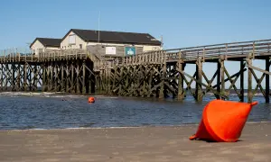 St. Peter-Ording an der Nordsee: Holzsteg mit Häusern, Bojen im Wasser und eine orangefarbene Boje am Strand.