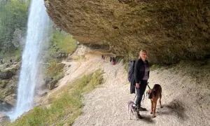 Frau mit zwei Hunden unter einem Felsvorsprung, während ein Wasserfall in der Nähe herabstürzt. Ein perfekter Ort, um die Natur zu genießen.