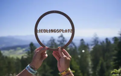 Hände halten ein Schild mit "LIEBLINGSPLATZ" vor einer malerischen Bergkulisse. Ein echter Geheimtipp im Bayerischen Wald.