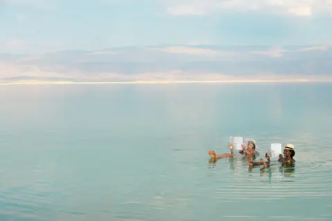 Zwei Frauen entspannen sich lesend im Toten Meer, dessen hoher Salzgehalt sie mühelos trägt. Der Meeresspiegel ist ruhig.