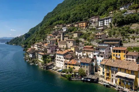 Bunte Häuser schmiegen sich an einen grünen Hang am Luganer See. Boote liegen am Ufer, die Sonne scheint.
