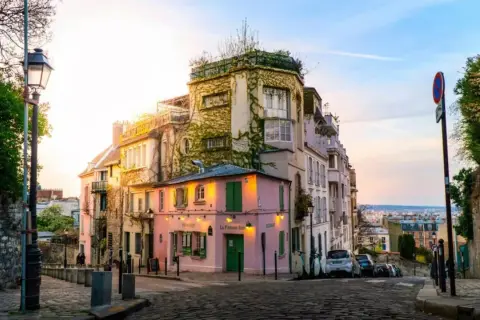 Malerische Gasse in Paris, perfekt für Sightseeing. Ein rosa Restaurant mit grünen Fensterläden und ein Mann mit Maske auf dem Kopfsteinpflaster.