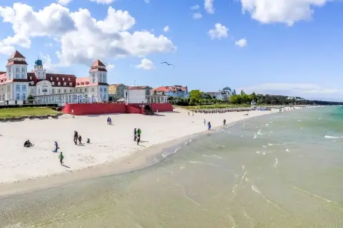 Ein sonniger Ostsee Strand mit Menschen, die spazieren gehen und Kinder spielen. Im Hintergrund ein prächtiges Gebäude im Bäderstil.