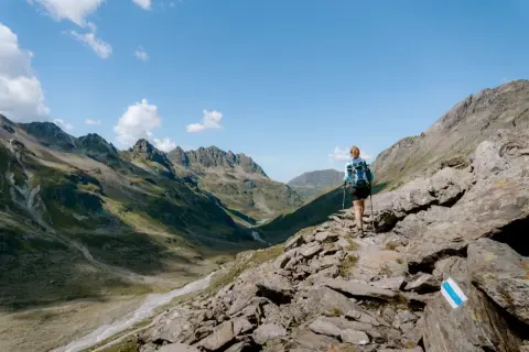 Frau mit Rucksack und Stöcken wandert auf felsigem Pfad im Montafon. Bergpanorama mit Tälern und Gipfeln.