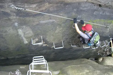 Ein Klettersteig im Elbsandsteingebirge: Eine Person sichert sich an einem Stahlseil, während sie über Felsstufen klettert.
