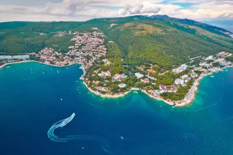 Blick auf eine Küstenstadt mit roten Dächern und grünen Hügeln, umgeben von türkisfarbenem Wasser. Ein Boot hinterlässt eine Spur im Meer. Ein echter **Istrien Geheimtipp**.