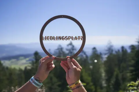 Hände halten ein Schild mit "LIEBLINGSPLATZ" vor einer malerischen Bergkulisse. Ein echter Geheimtipp im Bayerischen Wald.