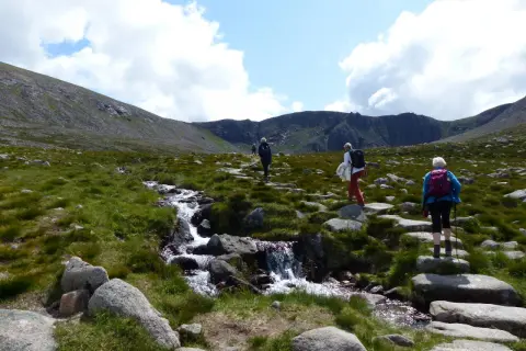 Drei Wanderer genießen das Cairngorms National Park Wandern entlang eines steinigen Pfades mit einem plätschernden Bach.