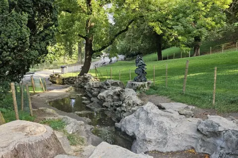 Ein kleiner Bach schlängelt sich durch den Parc des Buttes Chaumont, vorbei an Felsen und einer Skulptur. Grüne Hänge und Bäume säumen den Weg.