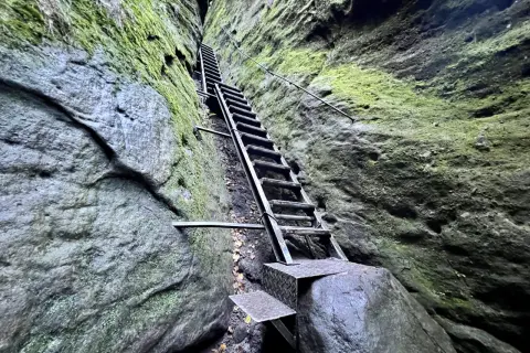 Steile Metalltreppe führt durch eine enge Felsspalte, Moos bedeckt die Wände. Eine leichte Schrammsteine Wanderung mit Ausblick.