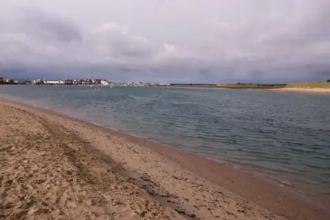 Sandiger Strand mit Blick auf eine Stadt und einen Yachthafen. Der Himmel ist bewölkt, aber das Wasser am Emden Strand ist ruhig.