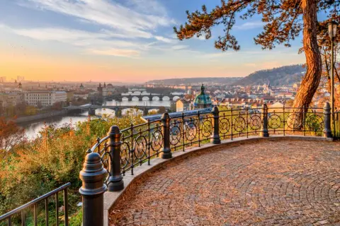 Prag im Herbst: Blick über die Moldau mit Brücken und Stadtkulisse bei Sonnenuntergang. Ein malerischer Ausblick von einem Aussichtspunkt mit gepflastertem Boden und schmiedeeisernem Geländer.