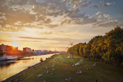 Sonnenuntergang an der Neckarwiese Mannheim: Menschen entspannen auf der Wiese, ein Schiff fährt auf dem Fluss, im Hintergrund Industrie und Bäume.