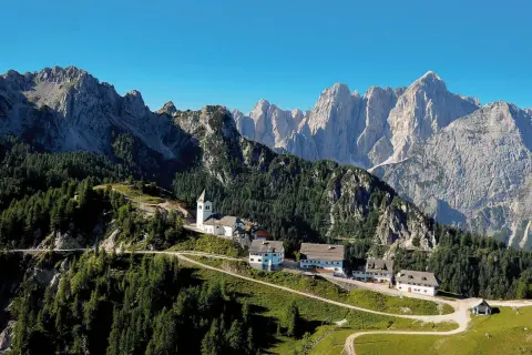 Alpe-Adria-Trail Erfahrungsbericht: Malerisches Bergdorf mit Kirche und Häusern, umgeben von Nadelwäldern und imposanten Gipfeln.