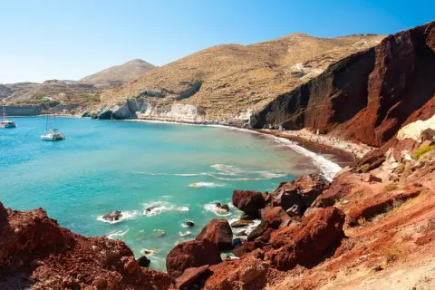 Der Rote Strand von Santorini mit türkisfarbenem Wasser, Segelbooten und Menschen, die sich auf dem roten Sand entspannen.