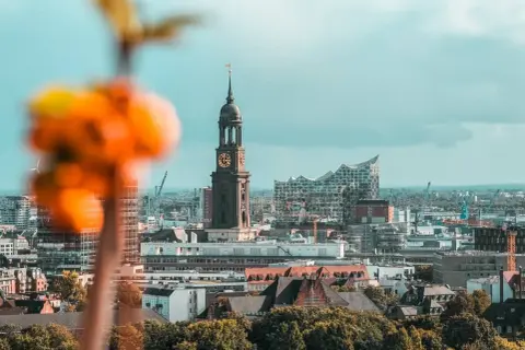 Blick auf Hamburg sehenswertes: Kirchturm St. Michaelis und Elbphilharmonie vor bewölktem Himmel. Im Vordergrund unscharfe orangefarbene Blüten.
