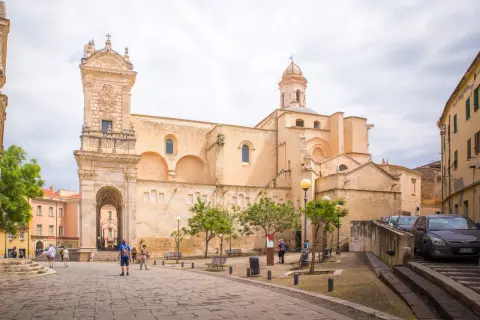 Ein sonniger Tag in der **Alghero Altstadt**. Menschen schlendern über den Platz vor einer imposanten Kirche mit einem verzierten Turm.