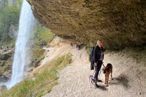 Frau mit zwei Hunden unter einem Felsvorsprung, während ein Wasserfall in der Nähe herabstürzt. Ein perfekter Ort, um die Natur zu genießen.