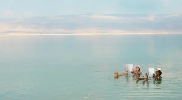 Zwei Frauen entspannen sich lesend im Toten Meer, dessen hoher Salzgehalt sie mühelos trägt. Der Meeresspiegel ist ruhig.