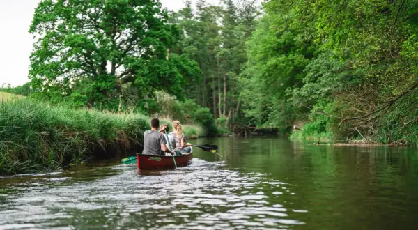Zwei Personen paddeln in einem Kanu durch einen Fluss, umgeben von üppiger grüner Natur. Ein friedlicher Moment im **Warnow Durchbruchstal**.