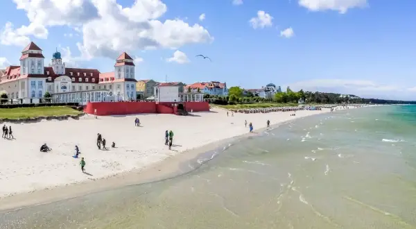 Ein sonniger Ostsee Strand mit Menschen, die spazieren gehen und Kinder spielen. Im Hintergrund ein prächtiges Gebäude im Bäderstil.