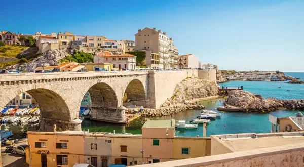 Bogenbrücke über einem Hafen mit Booten und Häusern, die sich an einen Hügel schmiegen. Die malerische marseille altstadt erstreckt sich bis zum türkisfarbenen Meer.