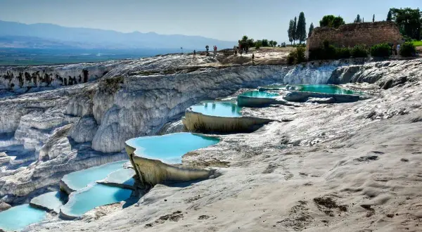 Travertinterrassen von Pamukkale heute: Türkisblaue Pools auf weißen Kalksteinterrassen, mit Besuchern im Hintergrund und antiken Ruinen.