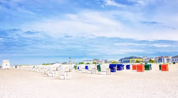 Strandkörbe in bunten Farben säumen den Sandstrand eines Ostseebads in Mecklenburg-Vorpommern. Im Hintergrund die Promenade mit Häusern und ein Leuchtturm.