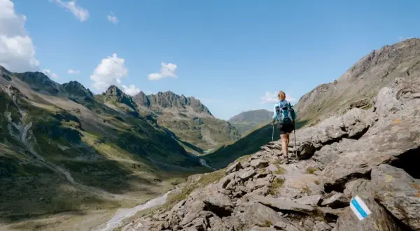 Frau mit Rucksack und Stöcken wandert auf felsigem Pfad im Montafon. Bergpanorama mit Tälern und Gipfeln.